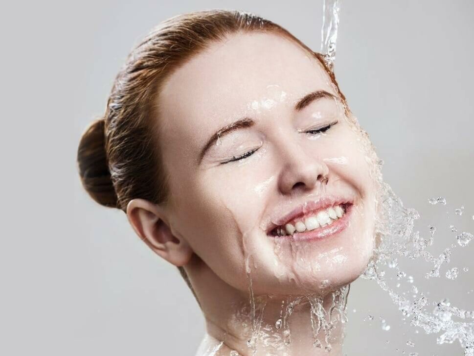 pH A person with their eyes closed and a joyful expression on their face tilts their head back under a stream of water, with droplets splashing around. Their hair is pulled back into a bun, and the background is a smooth, neutral shade.