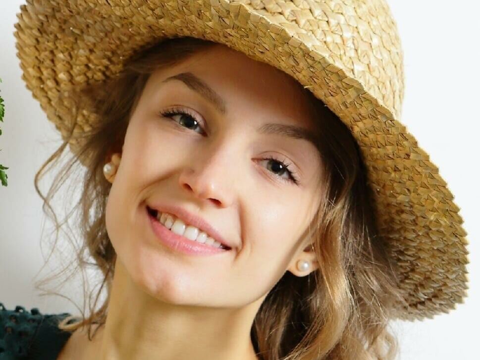 A woman with long, wavy hair smiles at the camera. She is wearing a wide-brimmed straw hat and small pearl earrings. The background is plain white, highlighting her cheerful expression and casual, summery appearance.