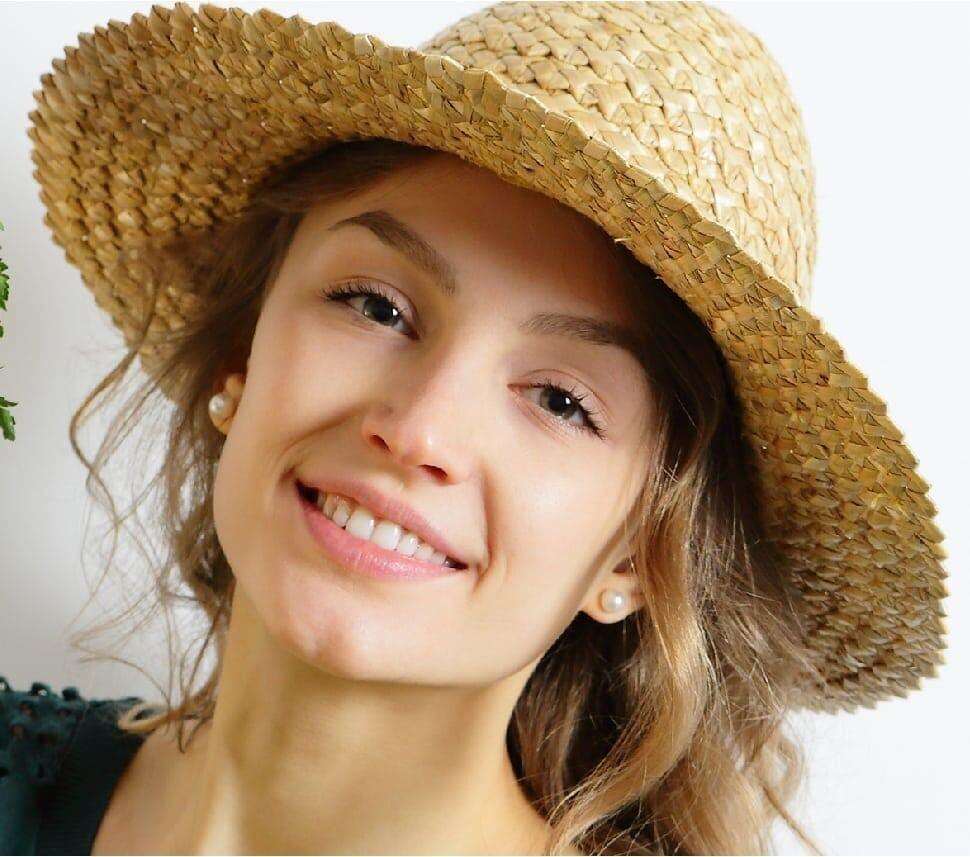A woman with long, wavy hair smiles at the camera. She is wearing a wide-brimmed straw hat and small pearl earrings. The background is plain white, highlighting her cheerful expression and casual, summery appearance.