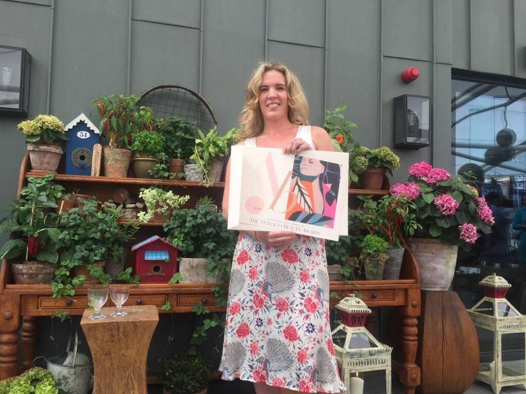 A woman in a floral dress is standing in front of a display of various potted plants and flowers. She is holding a colorful rectangular box. The setting appears to be a garden or a greenhouse with decorative items including birdhouses and lanterns.