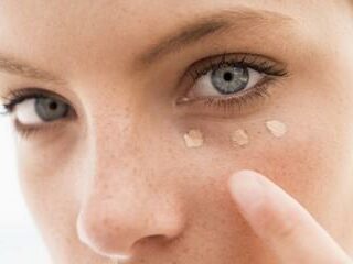 A close-up of a person with blue eyes applying three dots of light-colored concealer under their left eye using their finger. The skin appears natural with freckles, and the background is out of focus.