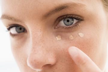 A close-up of a person with blue eyes applying three dots of light-colored concealer under their left eye using their finger. The skin appears natural with freckles, and the background is out of focus.