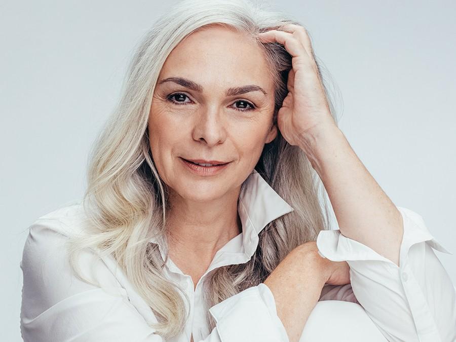 A middle-aged woman with long, gray hair and a calm expression rests her head on one hand. She is wearing a white shirt with the collar slightly popped and is posed against a plain white background.