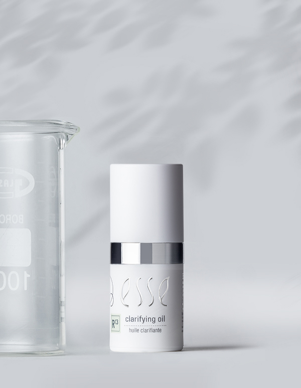 A white bottle of Esse Clarifying Serum is displayed on a light gray surface. The bottle has a silver band near its middle, and the text "P3 clarifying oil" is visible. A glass beaker is placed to the left of the bottle, partially obscured.