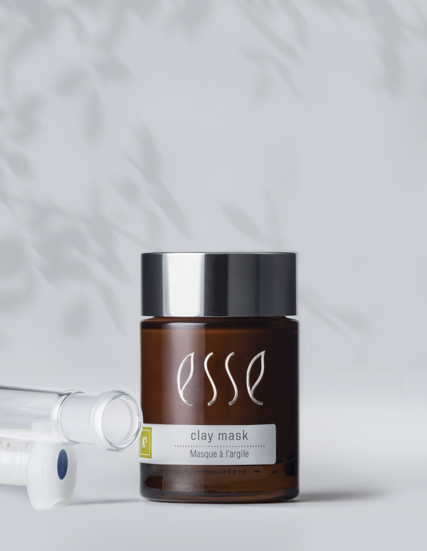 A brown glass jar of Esse Clay Mask with a black lid rests on a light gray surface. The label displays "Esse Clay Mask, Masque à l'argile." Beside the jar lies a clear glass dropper. Soft, blurry leaf shadows can be seen in the background.