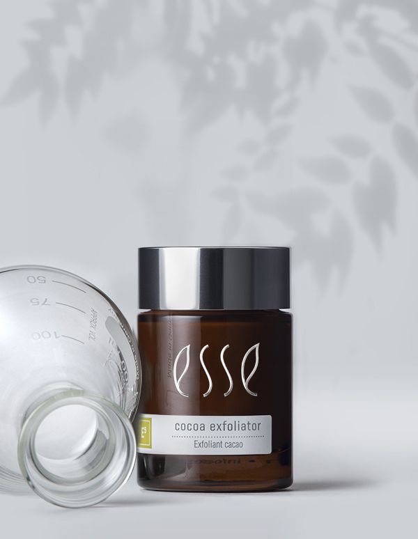 An Esse Cocoa Exfoliator jar, featuring a dark brown body with white text and a silver cap, stands upright beside a tipped-over transparent glass container. The light background with soft leaf shadows adds to the serene setting.
