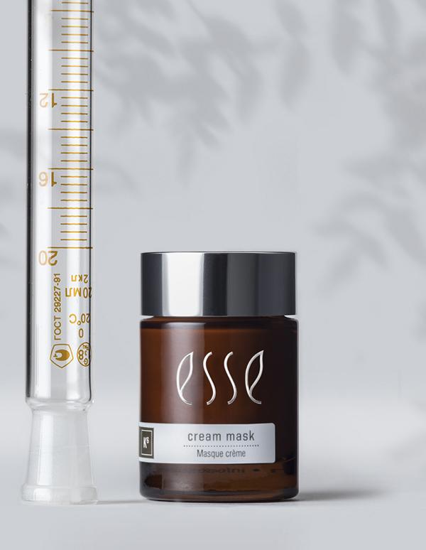 The Award-Winning Esse Cream Mask, housed in a brown glass jar with white and brown text and a silver lid, stands beside an upright glass tube marked with measurements. The background is light gray, accented by the shadow of leaves.