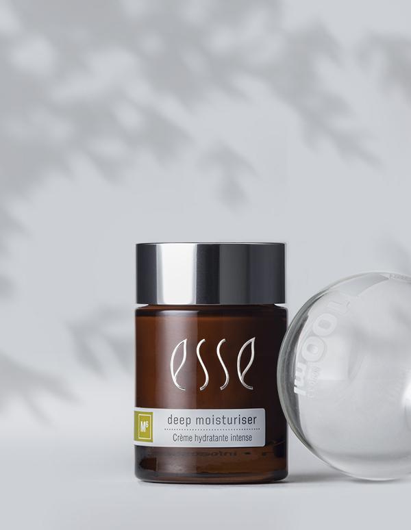 A jar of Esse Deep Moisturiser in brown, topped with a silver lid and a gray and white label, is placed against a light gray backdrop accented by leaf-shaped shadows. To the right, an open transparent sphere is partially visible.