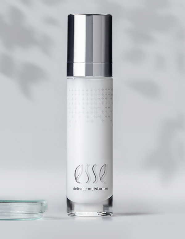 A sleek, cylindrical bottle of Esse Defence Moisturiser showcases a silver cap and minimalist design. The bottle contains white lotion and prominently displays the brand name "esse" along with the product name "defence moisturiser" on its front. The subtly blurred background enhances its elegant appearance.