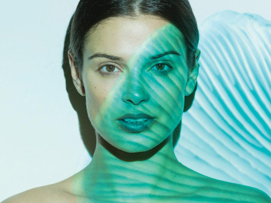 A woman with dark hair pulled back is partly illuminated by a green and blue striped light pattern. The stripes cast shadows on her face and body, creating an artistic effect. She gazes directly at the camera with a neutral expression.