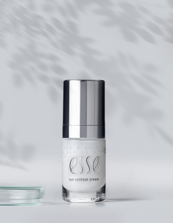 A small bottle of Esse Eye Contour Cream with a sleek silver cap is placed against a minimalist background with dappled shadow patterns. A glass object is partially visible in the lower left corner.
