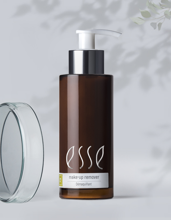 A brown bottle of Esse Make-up Remover with a white pump and a gray label stands against a light background with delicate shadowed leaves. The bottle's cap is also visible near the bottom left, and the product label includes the word "Démaquillant.