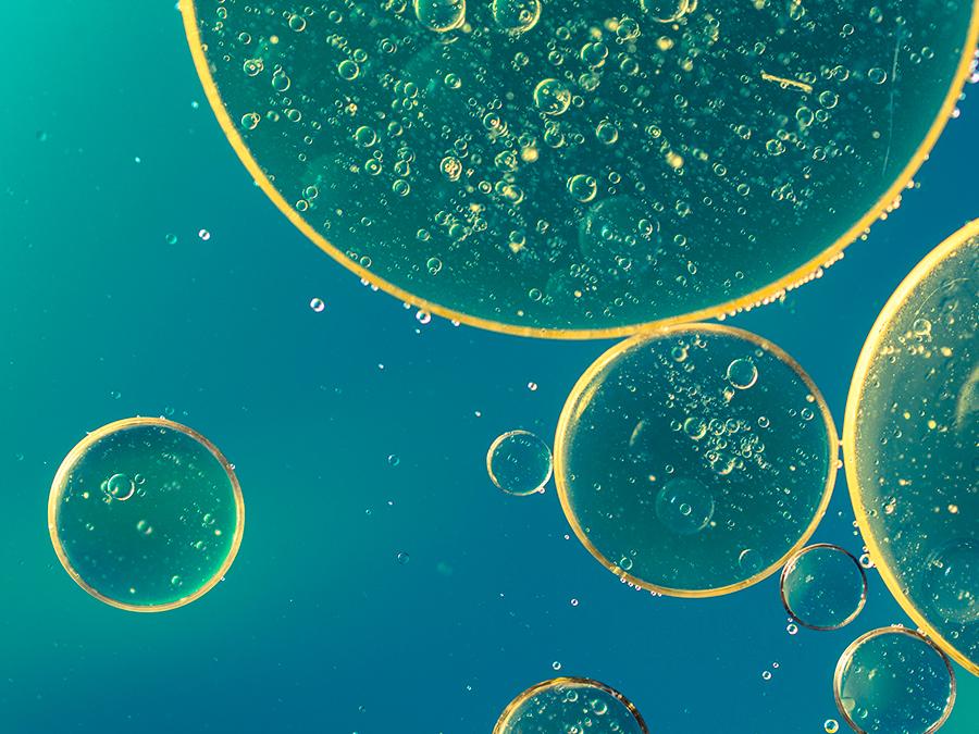 Hyaluronic-Acid Close-up of several translucent bubbles of various sizes floating in a teal liquid. Each bubble contains numerous tiny air pockets, creating a dotted texture. The light creates a slight glow around the edges of the bubbles, giving a whimsical effect.