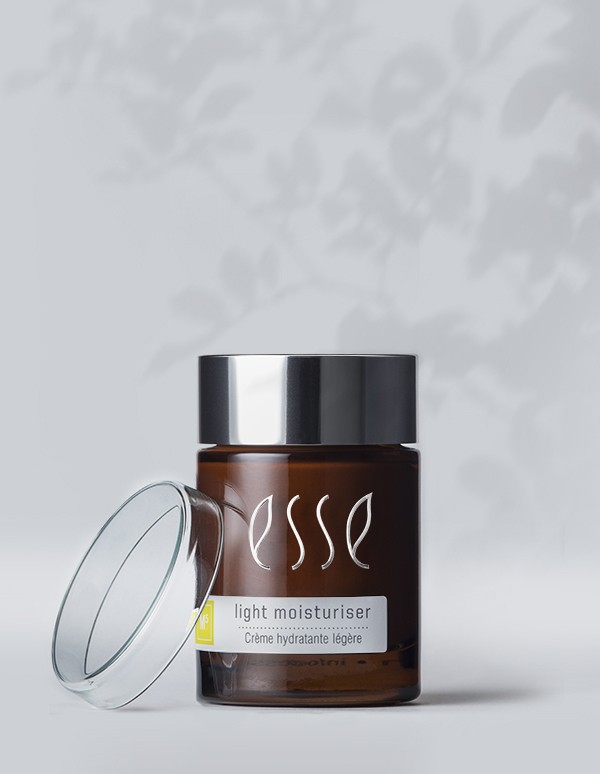 A jar of Esse Light Moisturiser with an open lid is displayed against a soft background with subtle plant shadows. The product label reads "Esse Light Moisturiser" and "Crème hydratante légère." The jar is dark brown with a silver cap.