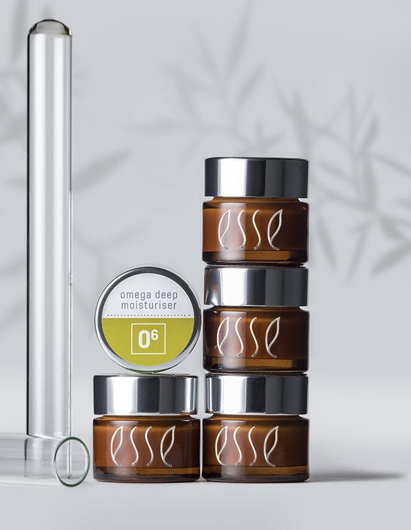 Four brown jars of Esse skincare products are stacked with a container labeled "Esse Omega Deep Moisturiser" on top. A test tube and the shadow of a plant branch are in the background on a light grey surface.