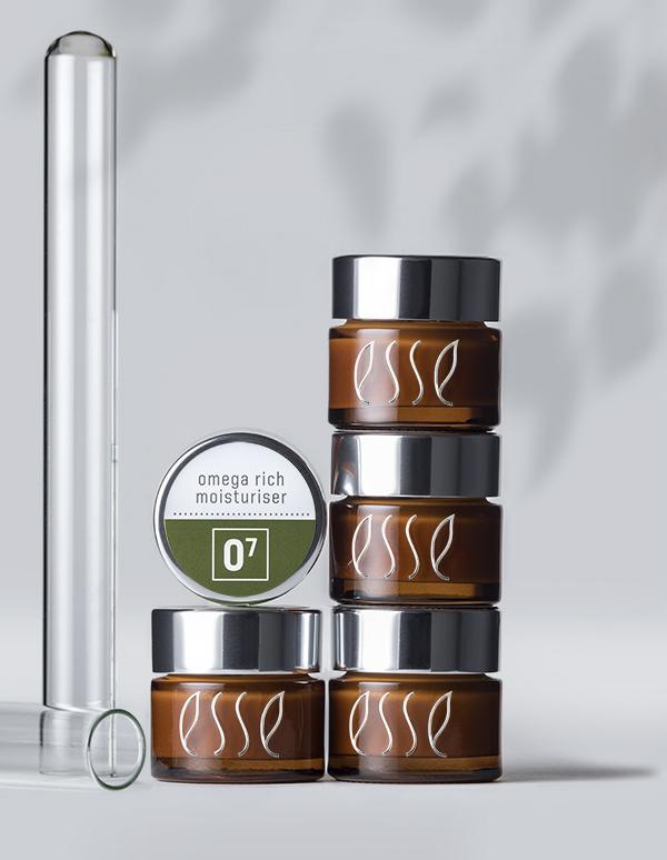 Multiple small amber jars with silver lids are stacked on top of each other, showcasing the brand name "esse" in white. A test tube leans against the stack, while a circular container labeled “Esse Omega Rich Moisturiser” is placed beside them.