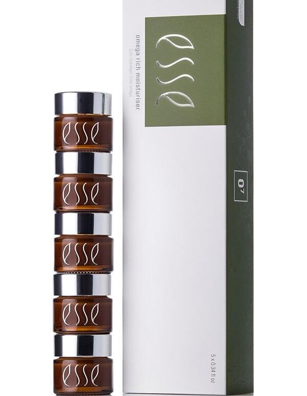 A stack of four brown "Esse Omega Rich Moisturiser" cosmetic jars with silver lids are placed beside a sleek white and green box featuring the same branding. The box stands upright on a white surface, showcasing the product packaging.