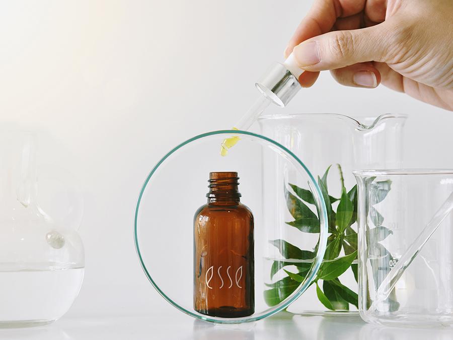 A hand is using a dropper to add liquid to a small brown glass bottle labeled "less." The bottle is inside a circular glass frame. Surrounding it are clear glass containers, one holding green leaves and another with a clear liquid, all set on a white surface.