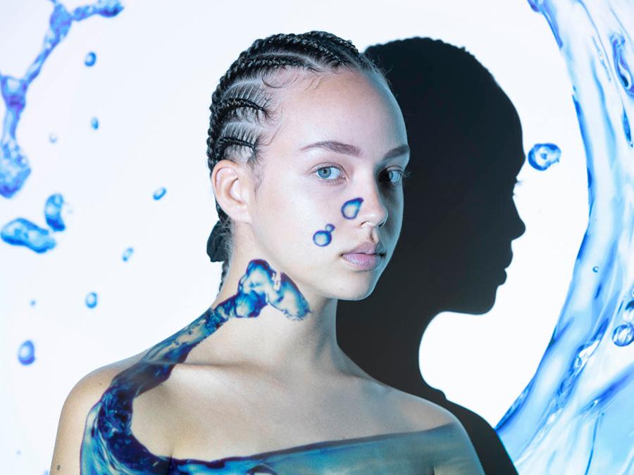 Probiotics-Moisturise A young person with cornrow braids stares forward against a light background with artistic water splashes and droplets. Their shadow is prominently visible on the wall behind them. The overall color scheme has cool blue and white tones.