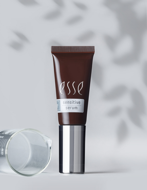 A tube of Esse Sensitive Serum stands upright against a light gray background with dappled shadows resembling leaves. The elegantly designed brown and silver packaging is paired with a clear, empty glass beaker resting on its side to the left.