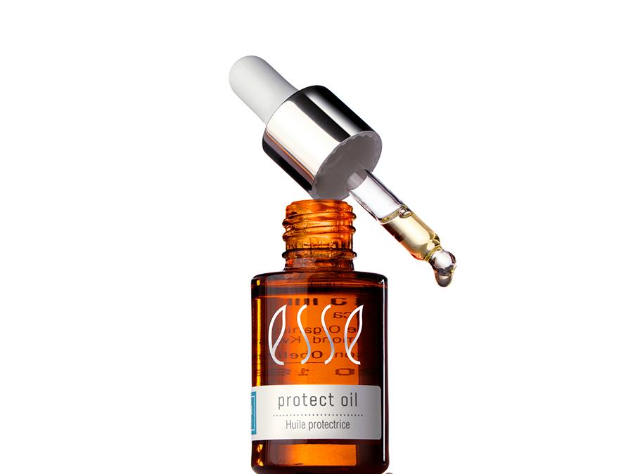 A brown glass bottle of protect oil with a label reading "Huile protectrice." The bottle is topped with a silver and white dropper, which is raised above the bottle, dispensing a drop of oil. The background is plain white.