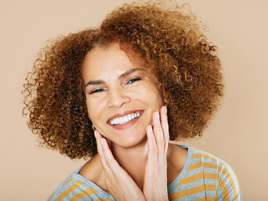 Anti-Ageing-blog-900-x-675px-2022 A smiling woman with curly hair touches her face with both hands. She is wearing a gray and yellow striped shirt, and stands against a beige background.
