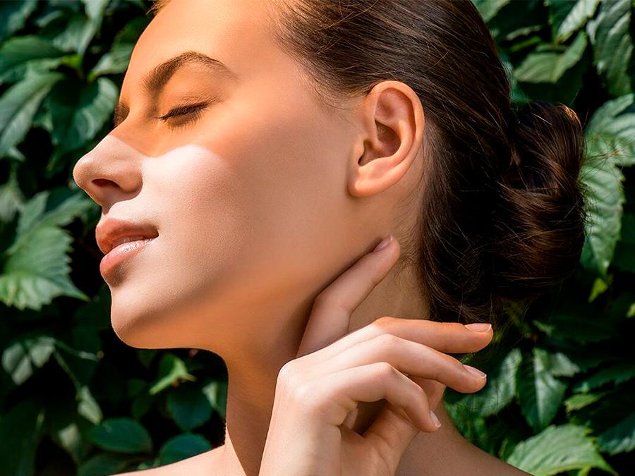 A person with smooth skin and closed eyes stands against a background of green leaves. They have their hair tied back in a bun and are lightly touching the side of their neck with their hand. Sunlight casts a warm glow on their face.