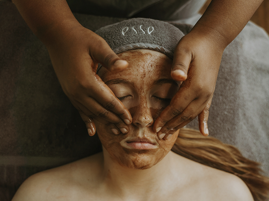 A person with long hair lies down with their eyes closed, receiving a facial treatment. They have a brown mask applied on their face, and a therapist’s hands are gently massaging it in. The person wears a gray headband embroidered with "esso.