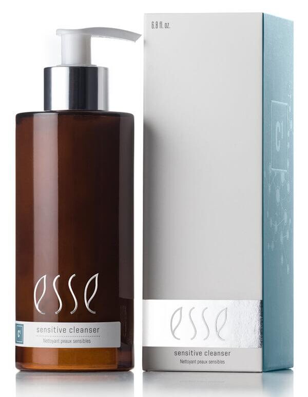 A bottle labeled "Esse Sensitive Cleanser" with a pump dispenser and brown in color is positioned next to a silver and white box that features the same branding. The bottle's label showcases white text, while the box has a metallic strip near the bottom providing product information.