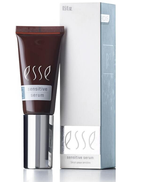 A sleek tube of Esse Sensitive Serum, in a sophisticated dark color, features a metallic silver cap and stands beside its light grey packaging box adorned with subtle decorative patterns. Both the box and the tube display the product name and a volume of 0.5 fl oz.