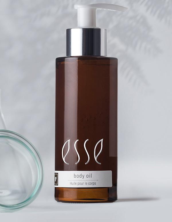 A brown bottle with a pump dispenser labeled "esse body oil - huile pour le corps." The bottle has sleek, minimalist design with a silver cap, set against a light background with a faint shadow of foliage.
