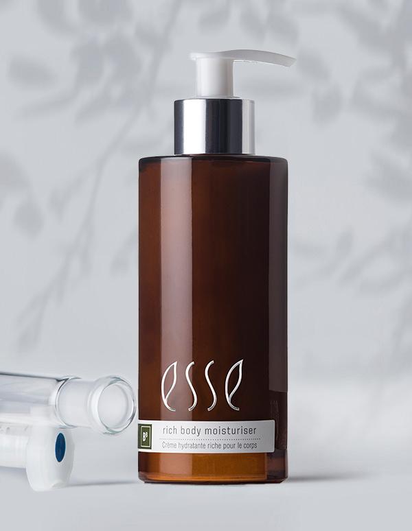 A brown bottle of Esse rich body moisturiser with a pump dispenser. The label has the brand name and product description in white text. The background is softly lit and includes blurred shadowed foliage, accompanied by a clear plastic cylindrical item near the bottle.