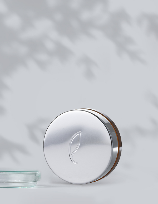 A round, silver tin labeled Esse Concealer with an embossed leaf design on the lid stands against a light gray background. The tin is partially open, revealing a hint of the concealer inside. Shadows of leaves create a subtle pattern on the wall in the background.
