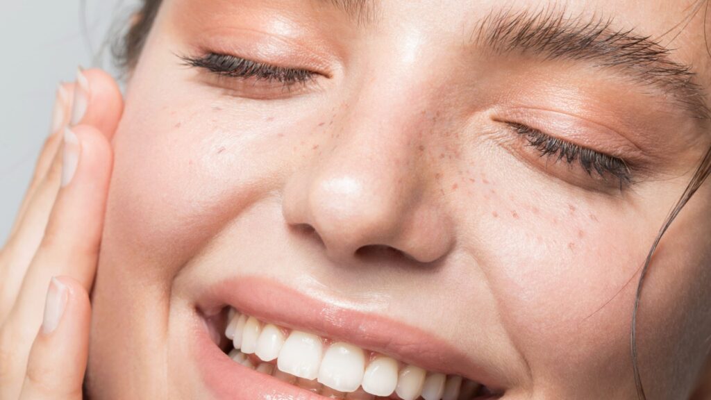 Close-up of a person with clear skin, freckles, and a bright smile. Their eyes are closed as they gently touch their face with one hand, showcasing radiant skin and natural beauty. The overall expression conveys happiness and satisfaction, emphasizing the effectiveness of the Esse Oily / Combination / Normal Trial / Travel Set.